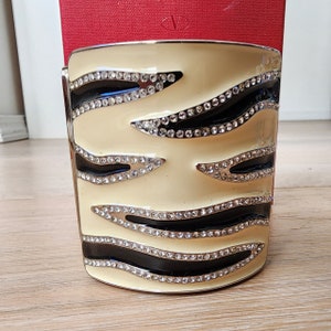 May include: A cream-colored metal bangle bracelet with a black and white zebra stripe design. The bracelet is adorned with clear rhinestones.