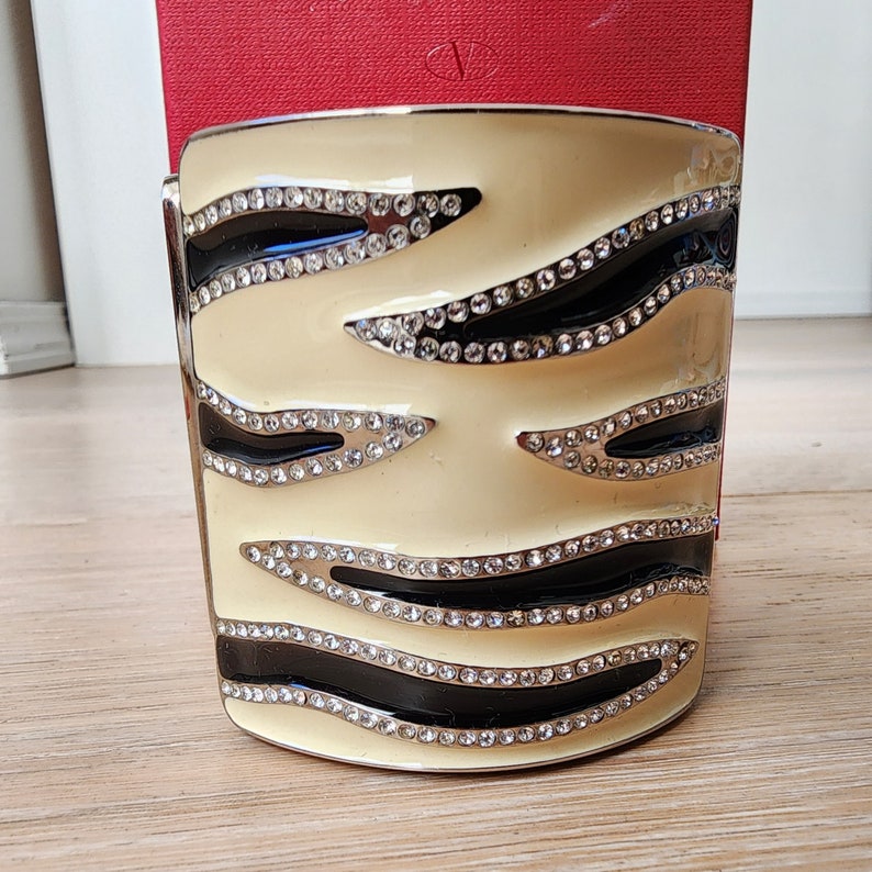 May include: A cream-colored metal bangle bracelet with a black and white zebra stripe design. The bracelet is adorned with clear rhinestones.