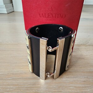 May include: Black leather cuff bracelet with silver accents and a yellow and white geometric design. The bracelet is made by Valentino Garavani.
