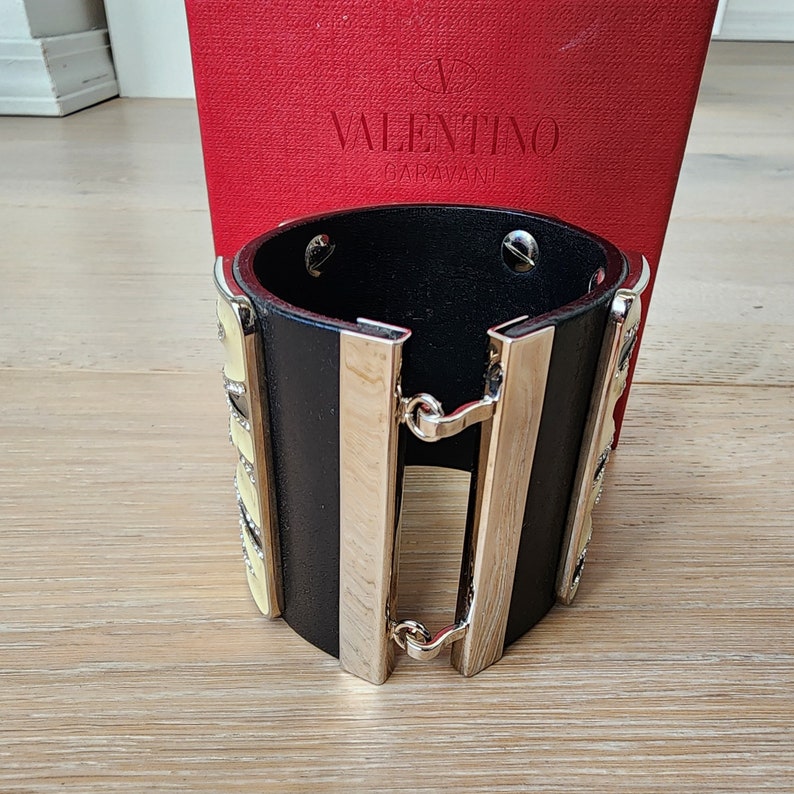 May include: Black leather cuff bracelet with silver accents and a yellow and white geometric design. The bracelet is made by Valentino Garavani.