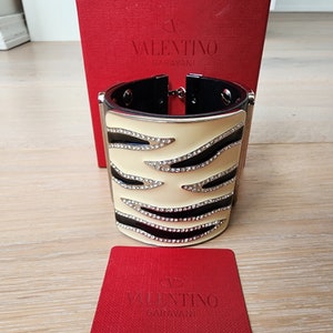 May include: A white and black zebra-print cuff bracelet with silver accents and rhinestones. The bracelet is shown with a red box with the Valentino Garavani logo.