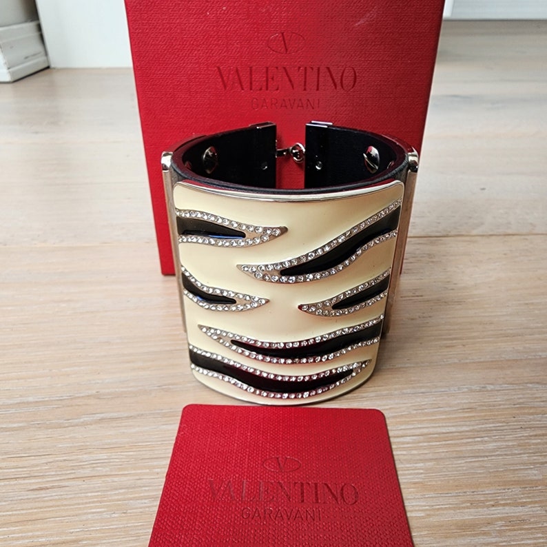 May include: A white and black zebra-print cuff bracelet with silver accents and rhinestones. The bracelet is shown with a red box with the Valentino Garavani logo.