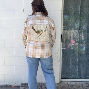 May include: A long-sleeve button-down shirt in a tan and white plaid pattern with a bleached effect. The back features a large, rectangular, quilted fabric patch displaying a detailed illustration of a bird perched on a branch.