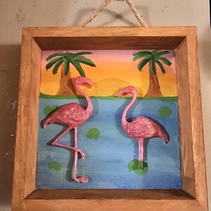 Peut inclure: Une image encadrée en bois avec une scène peinte d'un coucher de soleil sur un lac avec deux flamants roses. Les flamants roses sont en 3D et semblent être en plastique.