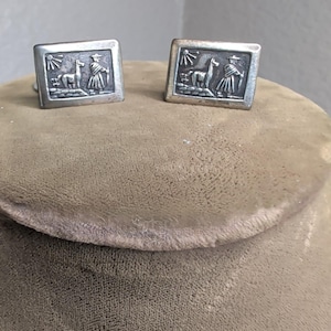 May include: A pair of rectangular silver-toned cufflinks featuring an embossed scene of a llama standing on a stone path next to a person wearing a wide-brimmed hat and a poncho, with a sunburst design in the upper left corner of each piece.