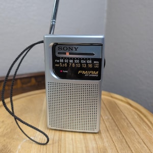 May include: A silver Sony ICF-S10MK2 portable FM/AM radio featuring a black display panel with frequency scales, a red tuning indicator, a speaker grille with small circular perforations, a retractable metal antenna, and a black wrist strap.
