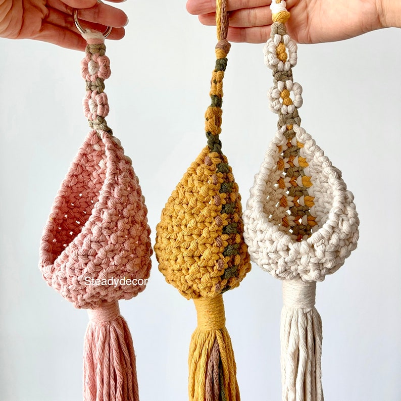 K&ouml;nnte beinhalten: Drei Makramee-H&auml;ngepflanzenhalter mit Quasten. Die Pflanzenhalter sind in rosa, gelb und wei&szlig;. Jeder Pflanzenhalter hat ein anderes Muster und Design.