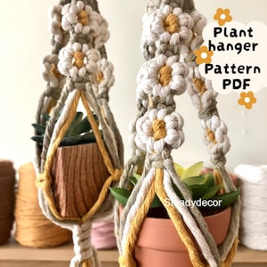 Può includere: Due portavasi in macramè con decorazioni a fiori di margherita. I portavasi sono realizzati con corda color crema, grigio e giallo e contengono vasi in terracotta con piante. L'immagine include il testo "Plant hanger Pattern PDF".