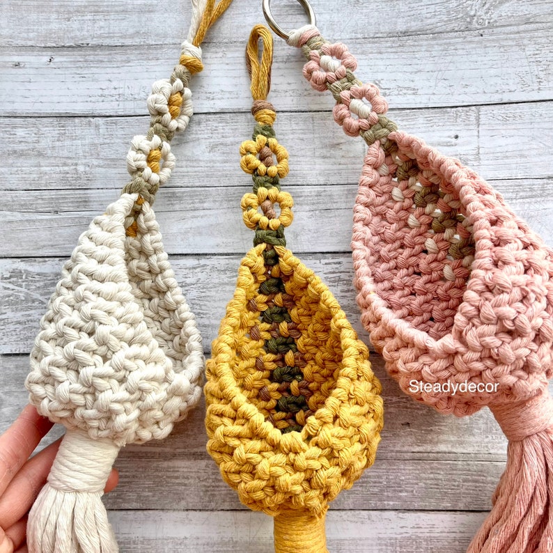 K&ouml;nnte beinhalten: Drei Makramee-Pflanzenh&auml;nger in verschiedenen Farben: Creme, Gelb und Rosa. Jeder H&auml;nger hat oben ein kleines Blumenmuster und unten einen Quasten. Der Text "Steadydecor" ist in der rechten unteren Ecke des Bildes sichtbar.