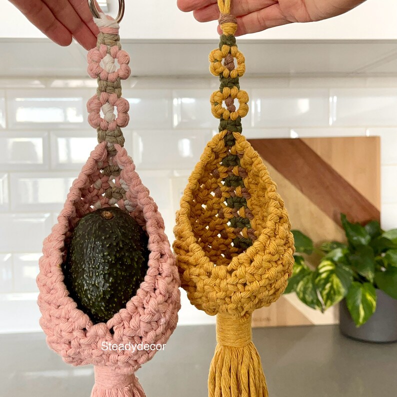 K&ouml;nnte beinhalten: Zwei Makramee-H&auml;ngepflanzen, eine rosa und eine gelbe, mit einer gr&uuml;nen Avocado in der rosa Pflanze. Die Pflanzen haben einen Quasten am Boden und sind mit kleinen wei&szlig;en und braunen Blumen verziert.