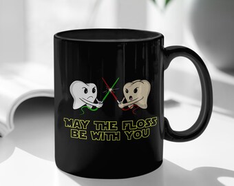 May include: Black coffee mug featuring a white and brown tooth design holding floss like lightsabers. The text reads "May the floss be with you".
