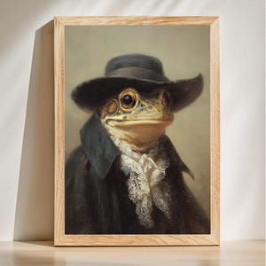 May include: A framed print featuring a frog dressed in a black hat and coat, with a white lace collar. The frog has large, expressive eyes and a serious expression. The artwork is set against a neutral background, with a wooden frame.