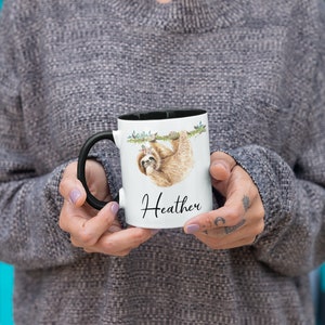 May include: White ceramic mug with a black rim and handle. The mug features a watercolour illustration of a sloth hanging from a branch with flowers. The text "Heather" is printed on the mug.
