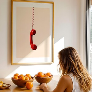 May include: A framed print of a red rotary phone hanging by a coiled cord against a light peach background.