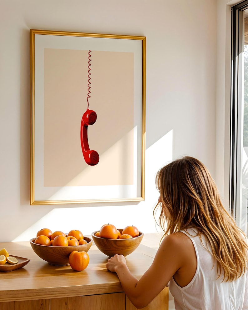 May include: A framed print of a red rotary phone hanging by a coiled cord against a light peach background.