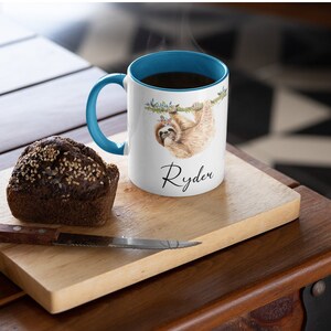 May include: White ceramic mug with a blue rim and handle. The mug features a watercolour illustration of a sloth hanging from a branch. The name "Ryden" is written in black cursive below the sloth.