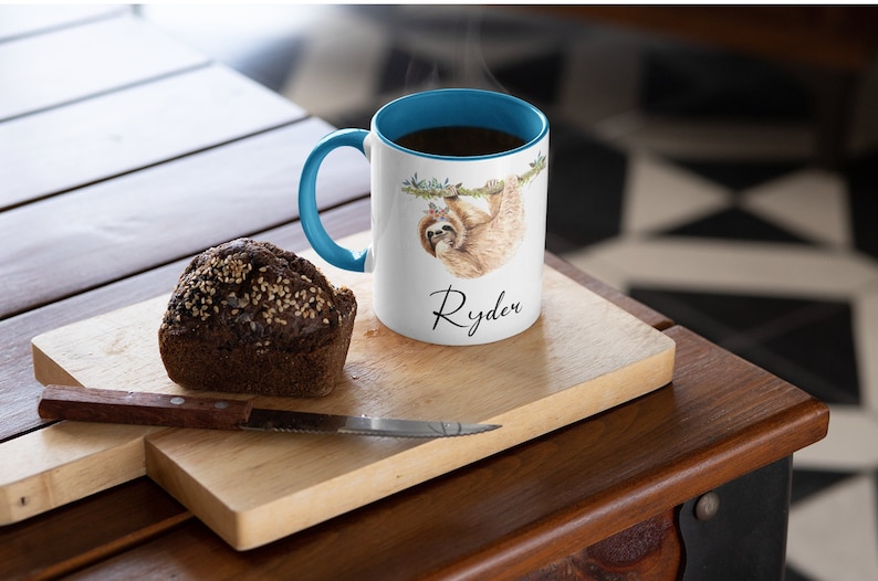 May include: White ceramic mug with a blue rim and handle. The mug features a watercolour illustration of a sloth hanging from a branch. The name "Ryden" is written in black cursive below the sloth.