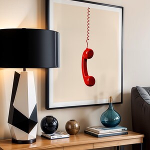 May include: A framed print of a red rotary phone hanging by a coiled cord against a beige background. The print is displayed on a wall above a wooden console table with a black and white geometric lamp, a black vase, a brown vase, and books.