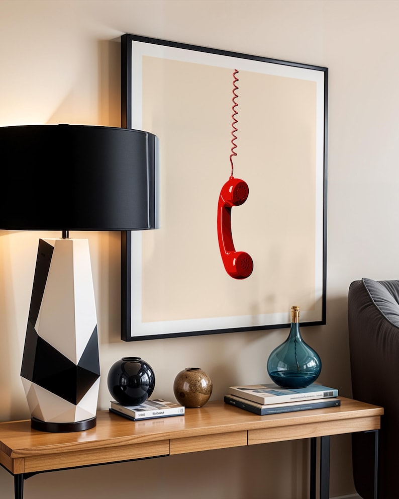 May include: A framed print of a red rotary phone hanging by a coiled cord against a beige background. The print is displayed on a wall above a wooden console table with a black and white geometric lamp, a black vase, a brown vase, and books.