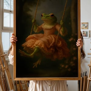 May include: A framed art print featuring a whimsical frog in a peach-colored dress, swinging on a rope swing. The frog has green skin and red eyes. The background is a dark forest scene with soft lighting.