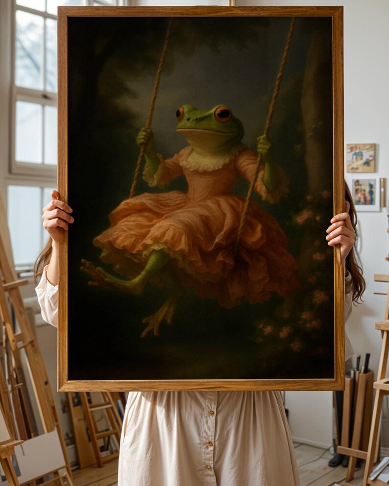 May include: A framed art print featuring a whimsical frog in a peach-colored dress, swinging on a rope swing. The frog has green skin and red eyes. The background is a dark forest scene with soft lighting.