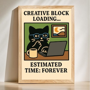 May include: A framed art print featuring a cartoon black cat wearing glasses and working on a laptop. The print includes the text "CREATIVE BLOCK LOADING..." and "ESTIMATED TIME: FOREVER". A mug and a small picture are also visible.