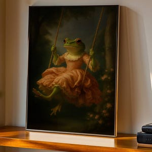 May include: A framed art print depicts a frog in a pink dress on a swing. The frog has green skin and red eyes. The swing is suspended from ropes against a dark, blurred background of trees and flowers.