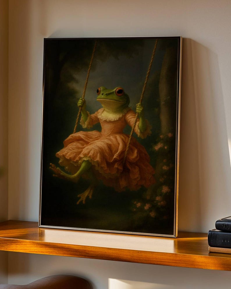 May include: A framed art print depicts a frog in a pink dress on a swing. The frog has green skin and red eyes. The swing is suspended from ropes against a dark, blurred background of trees and flowers.