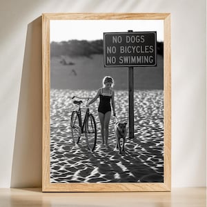 May include: Black and white photo of a girl walking a dog on a beach. The girl is wearing a swimsuit and holding a bicycle. A sign reads "No Dogs, No Bicycles, No Swimming."