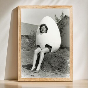 May include: Black and white framed print of a woman inside a large eggshell. The woman is sitting on a rock, wearing shorts and heels. The image has a vintage aesthetic, with a light wooden frame.