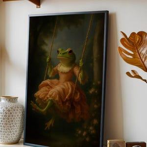 May include: Framed art print featuring a whimsical frog in a pink dress on a swing. The frog has green skin, red eyes, and is set against a dark, forest-like background. The artwork is framed in a black border.