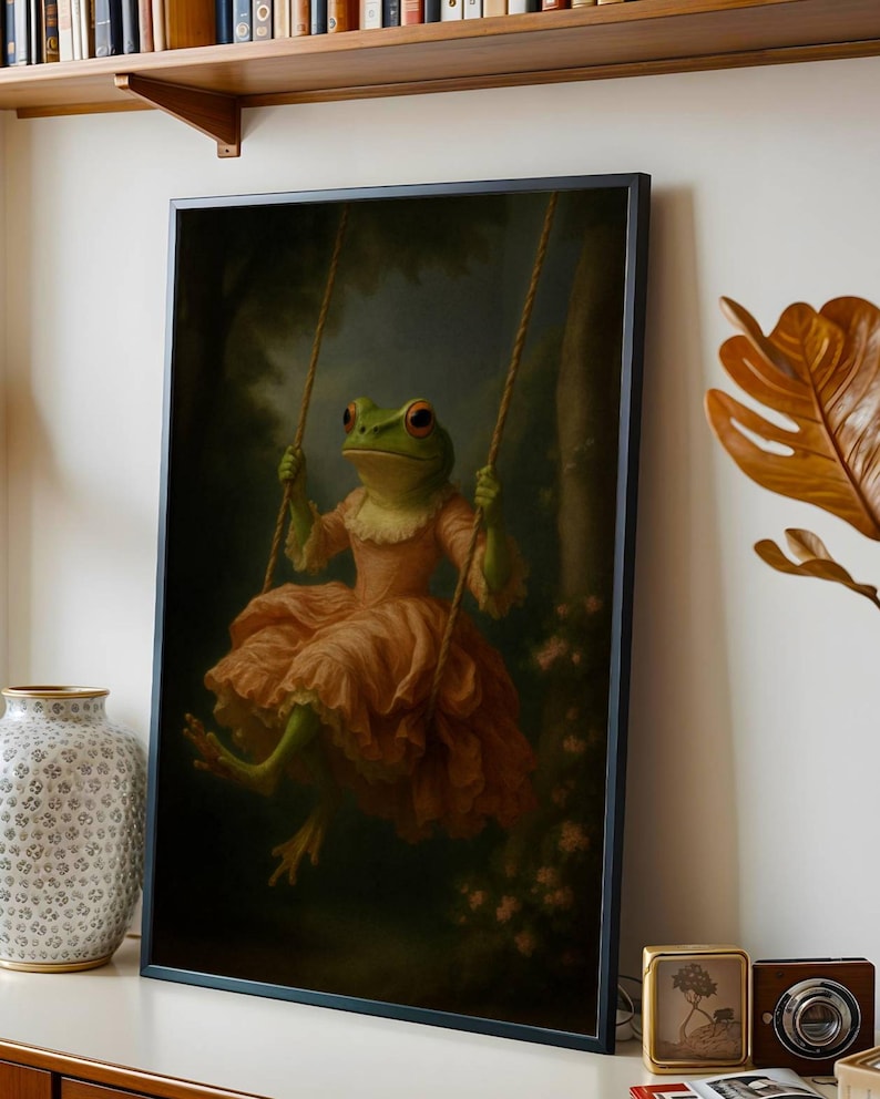 May include: Framed art print featuring a whimsical frog in a pink dress on a swing. The frog has green skin, red eyes, and is set against a dark, forest-like background. The artwork is framed in a black border.