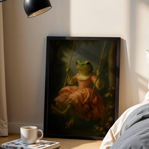 May include: A framed art print depicts a green frog in a peach-colored dress on a swing. The frog is in a forest setting with pink flowers. The artwork is displayed on a wooden floor next to a bed and a lamp.