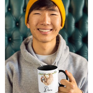 May include: A white ceramic mug with a black rim and handle. The mug features a watercolour illustration of a sloth hanging from a branch with a floral crown. The text "Dove" is printed in black below the sloth.