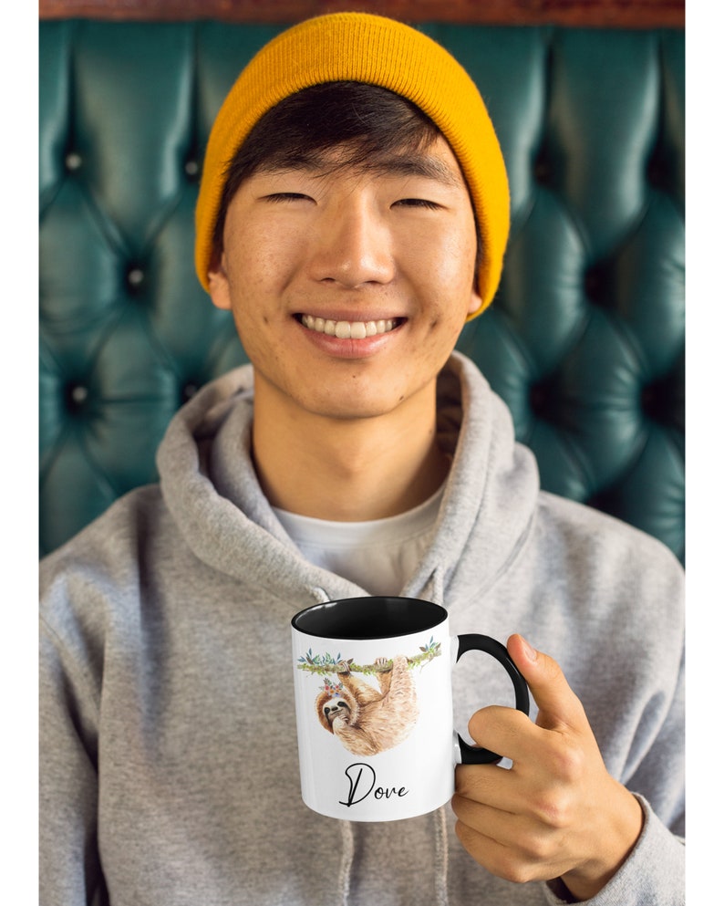 May include: A white ceramic mug with a black rim and handle. The mug features a watercolour illustration of a sloth hanging from a branch with a floral crown. The text "Dove" is printed in black below the sloth.