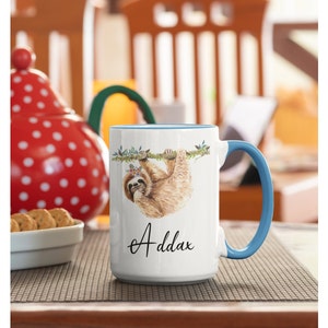 May include: White ceramic mug with a blue rim and handle. The mug features a watercolour illustration of a sloth hanging from a branch with flowers. The text "Addax" is printed in black cursive on the bottom of the mug.