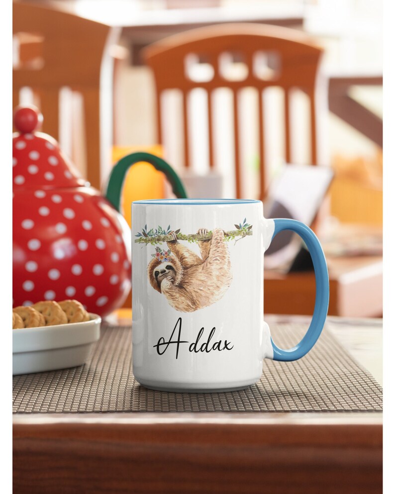 May include: White ceramic mug with a blue rim and handle. The mug features a watercolour illustration of a sloth hanging from a branch with flowers. The text "Addax" is printed in black cursive on the bottom of the mug.