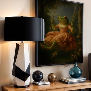 May include: A modern table lamp with a black lampshade and a geometric black and white base. A framed painting of a frog on a swing hangs on the wall. Decorative vases and books sit on a wooden table, creating a stylish interior design.