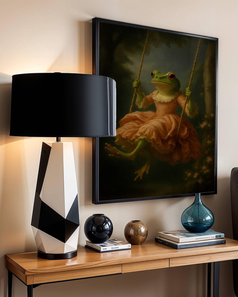 May include: A modern table lamp with a black lampshade and a geometric black and white base. A framed painting of a frog on a swing hangs on the wall. Decorative vases and books sit on a wooden table, creating a stylish interior design.
