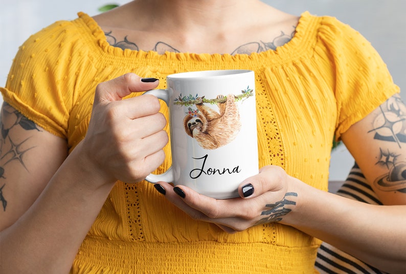 May include: White ceramic mug featuring a watercolour illustration of a sloth hanging from a branch. The sloth is wearing a flower crown. The mug is personalised with the name "Jonna" written in black cursive.