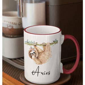 May include: White ceramic mug with a red rim and handle. The mug features a watercolour illustration of a sloth hanging from a branch with flowers in its hair. The text "Aries" is written in cursive below the sloth.