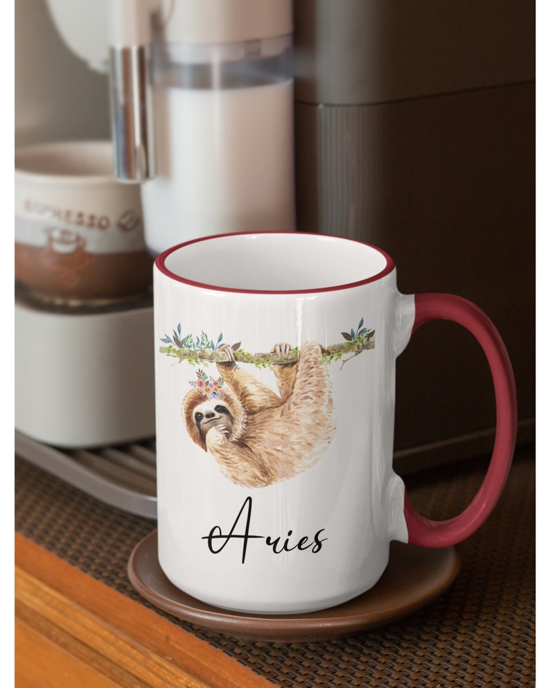 May include: White ceramic mug with a red rim and handle. The mug features a watercolour illustration of a sloth hanging from a branch with flowers in its hair. The text "Aries" is written in cursive below the sloth.