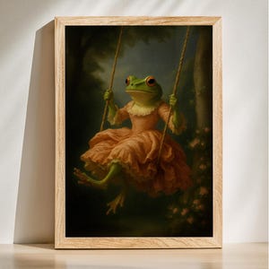 Puede incluir: Una impresión artística enmarcada que presenta una ilustración caprichosa de una rana en un columpio. La rana lleva un vestido color melocotón y está situada sobre un fondo oscuro, similar a un bosque. La obra de arte está en un marco de madera.