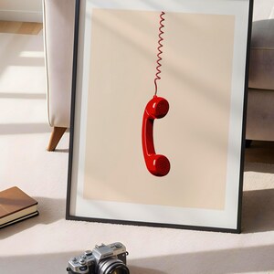 May include: A framed print of a red rotary phone handset hanging by a coiled cord against a light beige background.