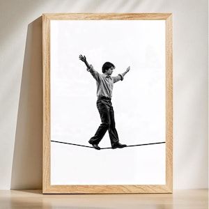 May include: Monochrome framed print showing a person balancing on a tightrope. The person is wearing a shirt and dark trousers, arms extended. The frame is made of light-coloured wood.