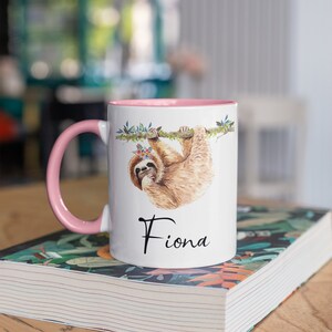 May include: A white ceramic mug with a pink rim and handle. The mug features a watercolour illustration of a sloth hanging from a branch with flowers. The name "Fiona" is printed in black below the sloth.
