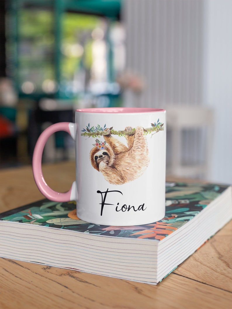 May include: A white ceramic mug with a pink rim and handle. The mug features a watercolour illustration of a sloth hanging from a branch with flowers. The name "Fiona" is printed in black below the sloth.