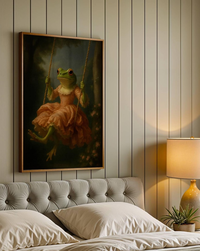 May include: A framed art print depicts a green frog in a pink dress on a swing. The frog has red eyes and is set against a dark forest background. The artwork is displayed above a bed with a tufted headboard.