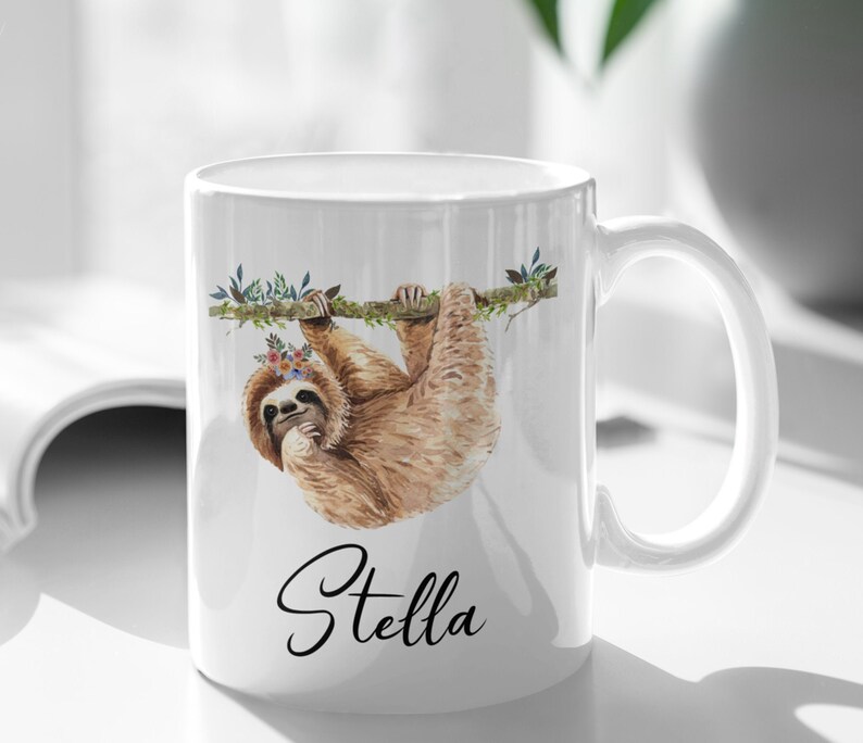 May include: White ceramic mug featuring a sloth hanging from a branch with flowers. The mug is personalised with the name "Stella" in black cursive.