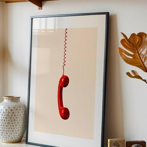 May include: A framed print of a red rotary phone receiver hanging by a coiled cord against a light beige background.